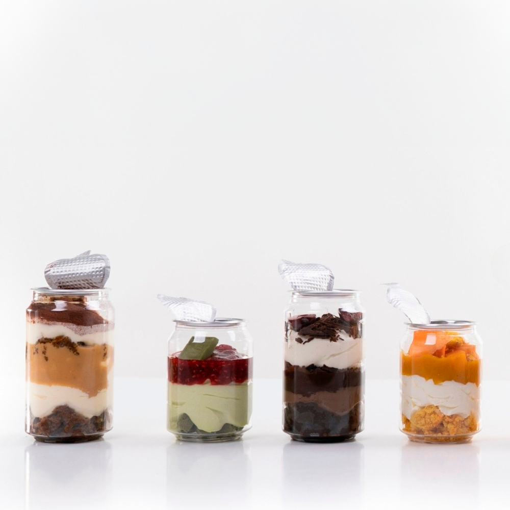 Cuatro tarros de vidrio con distintos postres por capas, como tiramisú, pistacho, chocolate y mango, con tapa metálica abierta sobre un fondo blanco.