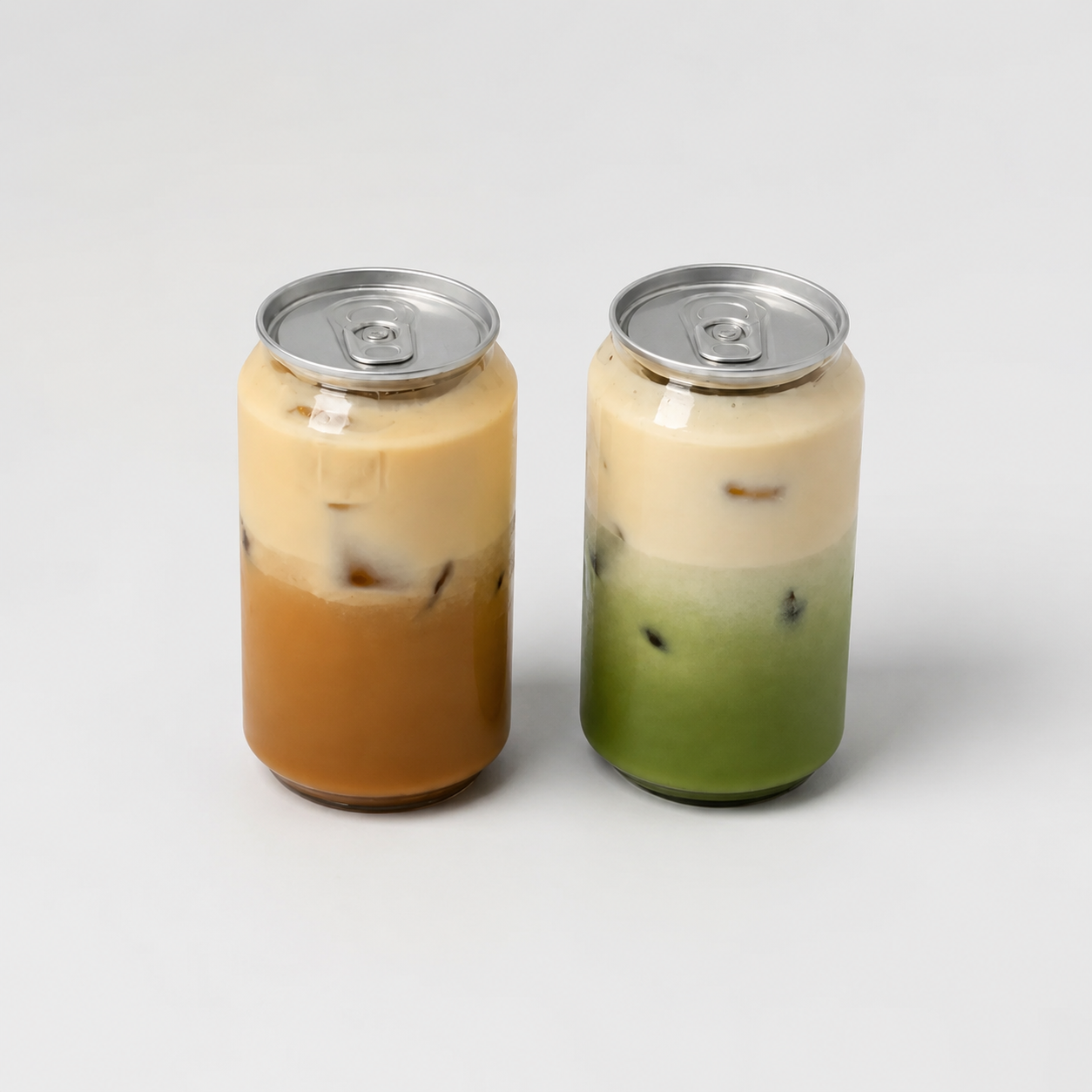 Dos latas transparentes con tapa plateada, una con café con leche helado y otra con matcha latte con hielo, sobre un fondo gris claro.